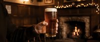 Man holding pint glass of beer in cozy pub interior with fireplace and warm lighting, perfect for beverage and hospitality marketing.