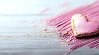 Pink heart-shaped macaron with gold glitter on pink feather against rustic wooden background, romantic food styling for Valentine's day and wedding concepts.