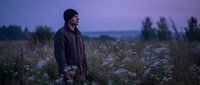 Young man with dark hair and light skin tone, wearing a dark jacket, standing alone in a wildflower meadow at dusk with purple twilight sky and distant mountains for contemplative lifestyle and nature themes.
