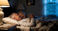 Senior couple embracing in bed at night, elderly man and woman sharing intimate moment in cozy bedroom with warm lamp light and moonlit window.