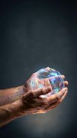 Hands gently holding glowing transparent sphere with rainbow reflections against dark background symbolizing protection, care and fragile dreams.