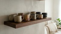Rustic ceramic mugs and vessels arranged on wooden shelf against neutral wall, showcasing handmade pottery collection for home decor and kitchenware styling.