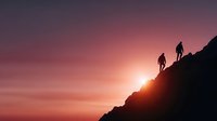 Two hikers climbing rocky mountain slope during dramatic sunset with pink and orange sky creating inspiring adventure silhouette for outdoor recreation concepts.