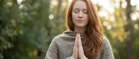 Young Caucasian woman with long red hair meditating outdoors in prayer pose wearing gray hoodie during golden hour sunset in peaceful forest setting.