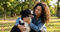 Young mixed-race woman with curly hair embracing black and white border collie dog in sunny park setting. Happy pet owner bonding moment outdoors.