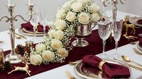 Elegant wedding table setting with white roses centerpiece, burgundy napkins, crystal glasses and golden candelabras for luxury celebration decor.