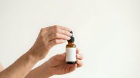 Male hands holding amber glass dropper bottle with white label against neutral background for skincare, cosmetics, or wellness product mockup.