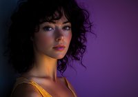 Young woman with curly dark hair in dramatic lighting against purple background, wearing yellow top, looking away with contemplative expression and natural makeup.