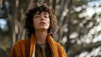 Young woman with curly hair wearing orange knitted cardigan outdoors in natural sunlight with trees in background for lifestyle and fashion content.