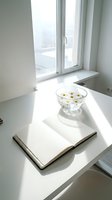 Minimalist white workspace with open book and glass bowl of daisies on windowsill in bright natural light for reading and relaxation.