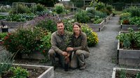 Two middle-aged adults, one male and one female, both of Caucasian ethnicity, crouch together in a community garden wearing work clothes, surrounded by raised beds of vegetables and flowering plants.
