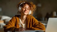 Young woman with curly hair wearing headphones and orange sweater enjoying music with eyes closed, relaxing at home workspace with laptop for lifestyle content.