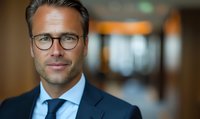 Confident mature Caucasian businessman wearing glasses and navy suit smiles warmly in modern office setting with soft bokeh background lighting.