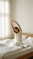 Young woman with dark hair and light skin tone stretching arms overhead while sitting on bed in bright morning light, wearing white casual shirt, promoting wellness, relaxation and healthy sleep routines.