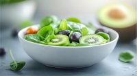 Fresh green spinach salad with sliced kiwi, blueberries and baby carrots in white ceramic bowl, healthy eating concept on gray background with avocado.