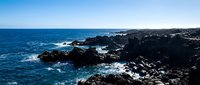 Dramatic rocky coastline with dark volcanic rocks meeting turquoise ocean waves under clear blue sky, suitable for travel and nature backgrounds.