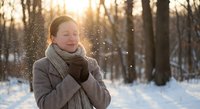 Young Asian woman enjoying winter snowfall in forest at golden hour sunset with warm scarf and coat feeling joyful and peaceful in snowy woodland landscape.