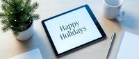 Tablet on wooden desk displaying Happy Holidays message, with Christmas tree, coffee mug, and pen, festive holiday greeting concept.