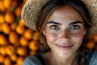 Young woman with bright blue eyes and warm smile wearing straw hat against blurred orange citrus fruit background, natural summer portrait capturing genuine happiness.