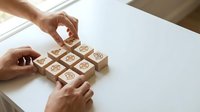 Hands arranging wooden blocks with engraved business and communication icons on a white table, symbolizing strategy and teamwork.