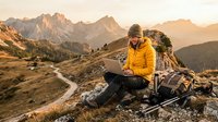 Male hiker in yellow jacket using laptop on mountain peak during golden hour sunrise with dramatic alpine landscape and winding trail below.