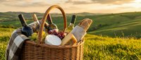 Wicker picnic basket filled with wine bottles, bread, cheese and checkered cloth on sunny hillside meadow with rolling green landscape.
