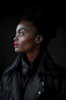 Young African American woman with red lipstick and hair bun in black jacket, photographed in dramatic side profile against dark background. Artistic portrait with moody lighting.
