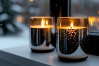 Glowing candles in black glass holders on windowsill create cozy atmosphere against snowy winter background, warm candlelight illuminates dark evening.