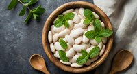 White beans in wooden bowl with fresh mint leaves on dark stone background for healthy cooking and nutrition concepts.