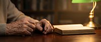 Elderly hands turning pages of open book on wooden desk with vintage brass lamp creating warm atmospheric reading environment for literature study.