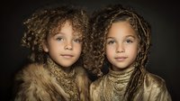 Two young mixed-race girls with curly hair and braids wearing cozy winter scarves and fur-trimmed jackets against dark background for fashion portrait.
