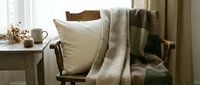 Cozy wooden armchair with cream pillow and beige throw blanket in warm sunlit room. Rustic home decor with coffee cup and dried flowers on side table.