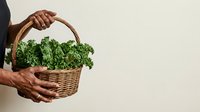Farmer with dark skin tone holding wicker basket filled with fresh green kale leaves against neutral background for organic produce and agriculture concepts.