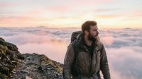 Bearded male hiker in winter gear stands on rocky mountain peak above sea of clouds at sunrise, contemplating vast landscape during outdoor adventure.