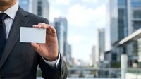 Businessman in dark suit and tie holding blank white business card with urban skyline and modern office buildings in background.