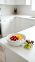 Healthy breakfast bowl with oatmeal, fresh raspberries, yellow mango slices, and granola in modern white kitchen with grapes on counter.
