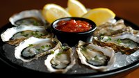 Fresh raw oysters served on crushed ice with lemon wedges and spicy cocktail sauce, garnished with herbs on dark plate, restaurant appetizer presentation.