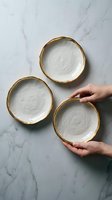 Three ceramic bowls with gold-trimmed rims arranged on marble surface, two held by hands with light skin tone. Suitable for tableware, home decor, and dining concepts.