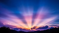 Dramatic sunset rays pierce through clouds over mountain silhouette, creating vibrant purple and orange sky with radiating crepuscular beams. Nature landscape background.
