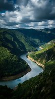 Aerial view of a winding river cutting through lush green forested mountains under dramatic cloudy skies, suitable for travel and nature editorial projects.