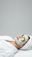 Woman relaxing with green cucumber facial mask and slices on eyes wearing white bathrobe for spa skincare treatment and wellness routine.