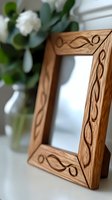 Handcrafted wooden photo frame with carved vine pattern detail, shown at angle with soft focus white flowers in glass vase background. Natural wood grain texture.