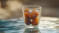 Fresh dates soaking in clear water glass with natural sunlight casting soft shadows on blue surface, healthy organic dried fruit preparation.