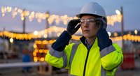 Asian female construction worker in safety gear talking on phone at illuminated industrial site during evening hours with bokeh lights in background.