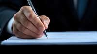 Close-up of businessman's hand signing document with pen in dark office setting, representing corporate agreement, contract, or legal paperwork.