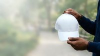 Male construction worker with dark skin tone holding white safety helmet in both hands outdoors on blurred site background for occupational safety and workplace protection concepts.