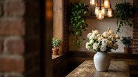 Cozy rustic interior with white roses in ceramic vase on wooden table, exposed brick wall, hanging Edison bulbs and green plants creating warm atmospheric lighting.