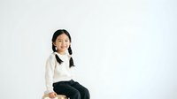 Young Asian woman with long black hair wearing a cream-colored sweater and black pants, sitting against a white background for portrait and lifestyle use.