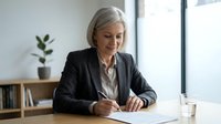 Senior businesswoman with gray hair writing documents at office desk, professional executive working on paperwork in modern workplace.