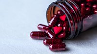 Red glittering capsules spilling from glass bottle on white surface, pharmaceutical supplements with sparkly coating in deep burgundy color. Medical concept.